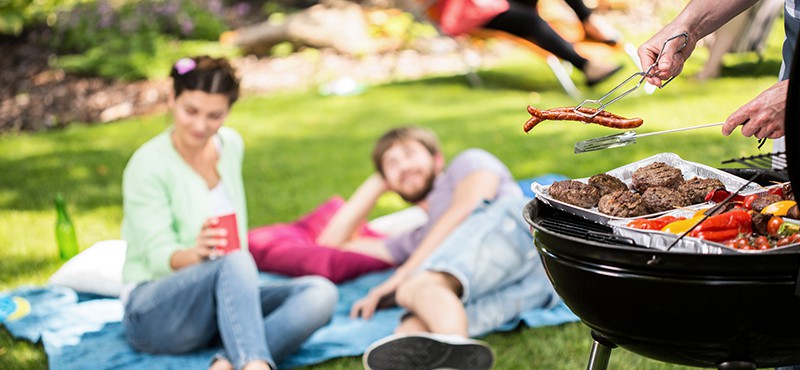 two people resting on picnic blanket in grass with someone grilling in foreground